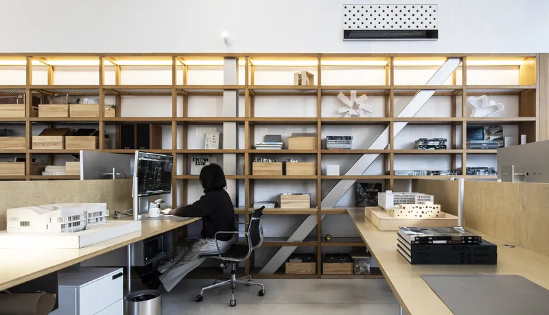 Light wood shelving wall with integrated desk and architectural models under warm linear lighting