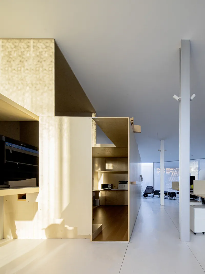 View through an angular plywood-clad passage opening to a white gallery space with columns beyond