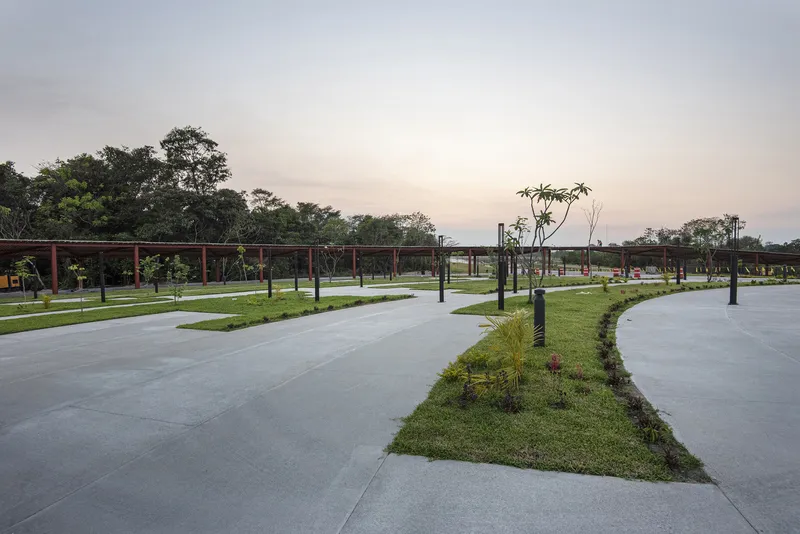 Paved plaza with planted beds and young trees at dusk beneath the red colonnade