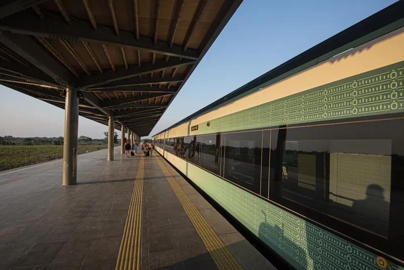 Covered platform with timber columns and yellow tactile paving alongside green perforated facade at dusk