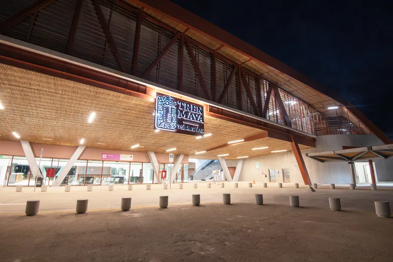 Night view of the arrival plaza with angled white columns supporting the timber-clad canopy and woven ceiling panels