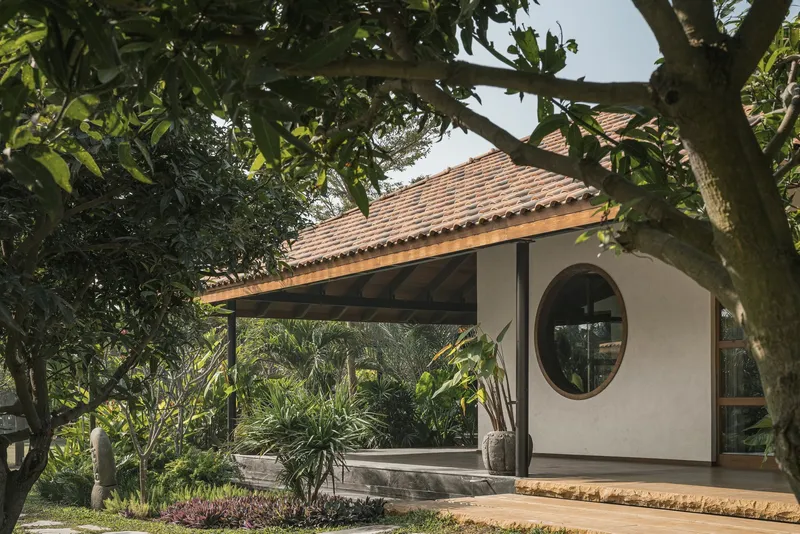 White stucco pavilion with circular window and clay tile roof beneath a canopy of mature trees