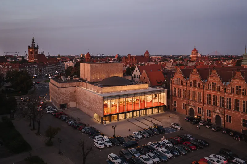 Elevated view of the illuminated glass entry volume adjacent to historic brick gabled buildings at dusk