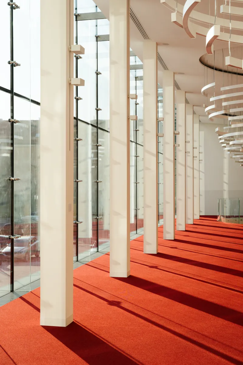 White tapered columns aligned along the glass facade with red stepped flooring in afternoon light
