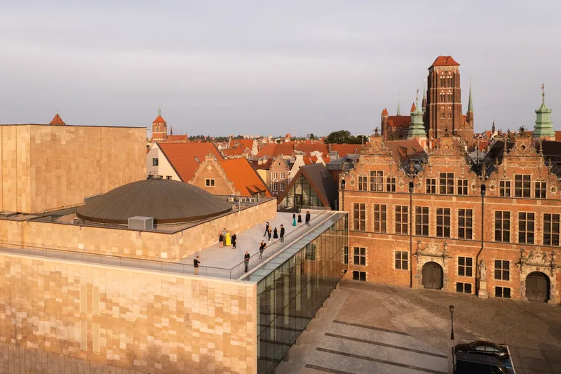 Rooftop terrace with visitors walking beside the glazed volume overlooking the historic tiled roofscape