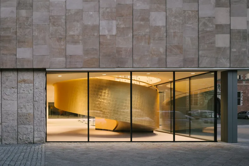 Street-level glazed entrance with illuminated curved interior volume behind vertical stone cladding at dusk