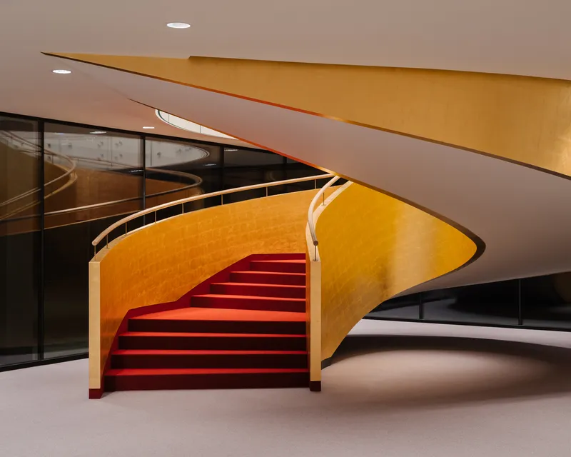 Curving staircase with red runner ascending between gold-leafed walls and glass railings