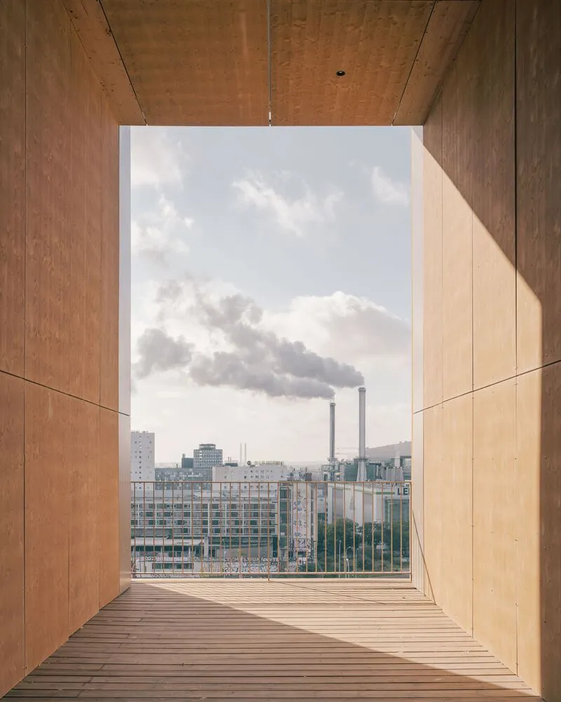 Timber-lined window opening framing industrial buildings and a smoking chimney stack beyond