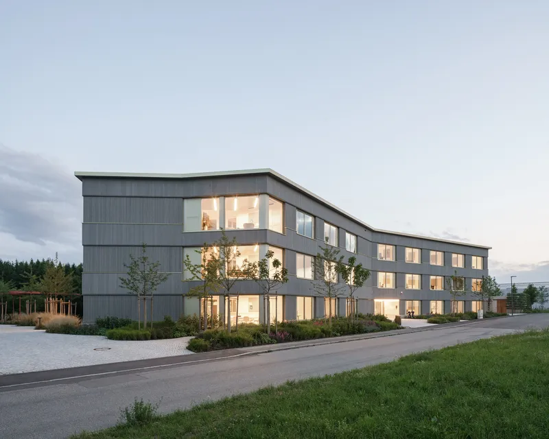 Three-story facade with grey ribbed cladding and illuminated windows at dusk
