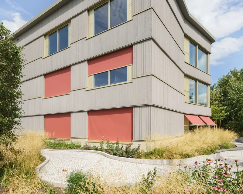 Corner facade with pale ribbed panels and coral-colored awnings above naturalized plantings