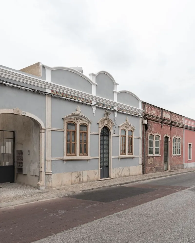 Angled street view showing the renovated pale blue facade with arched parapet next to weathered red neighbor