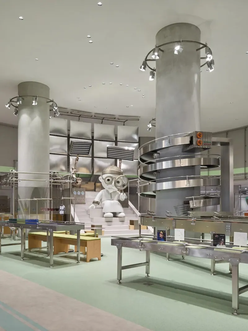 Concrete columns with ring-mounted spotlights above steel conveyors and white robotic figures on display platforms