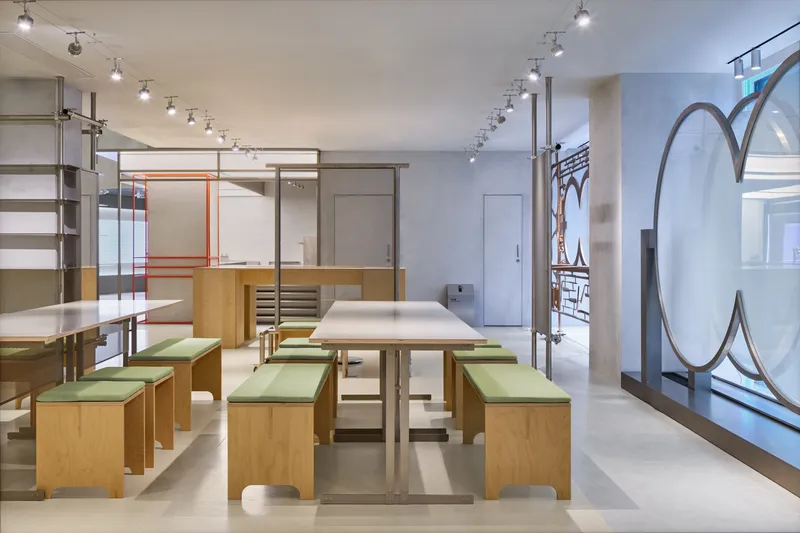 Light wood benches with green cushions arranged around steel table under track lighting and storefront glazing