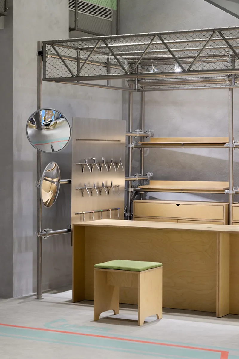 Wood counter with green upholstered stool beside steel truss framework holding suspended tools and round mirrors