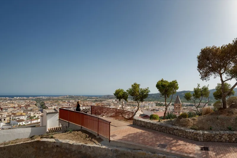 Elevated terrace with corrugated metal railing and brick paving overlooking a hillside town and distant sea