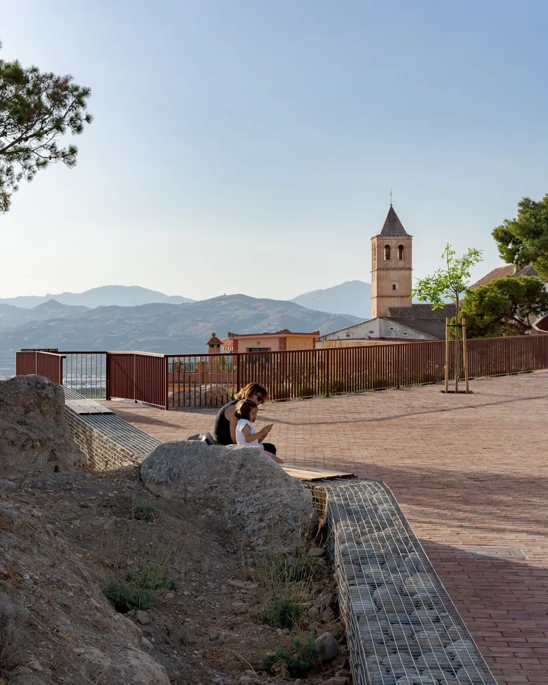 Brick-paved promenade with woman seated on a rock outcrop beside gabion retaining wall and metal balustrade