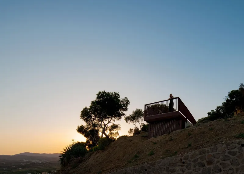 Brick observation platform with metal railing on hillside silhouetted against sunset with solitary figure