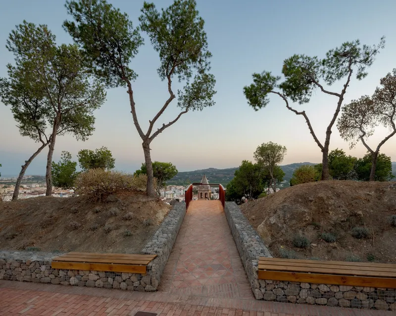 Brick paved pathway between gabion wall benches flanked by trees leading downhill at twilight