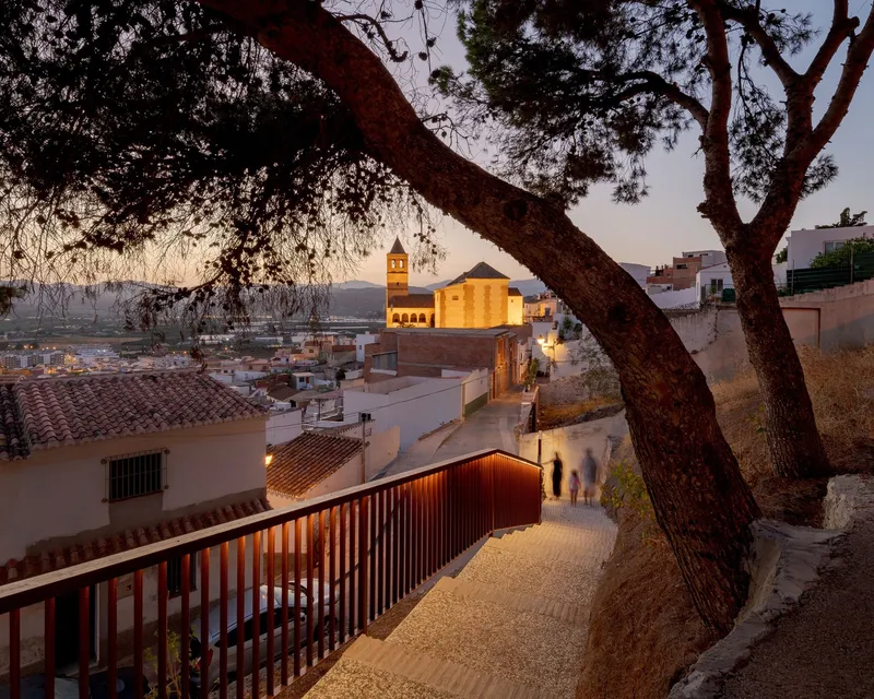Gravel pathway descending between red metal balustrade and church tower framed by overhanging pine branches