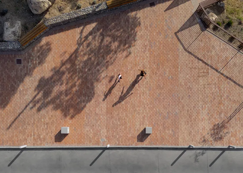 Aerial view of terracotta brick plaza with two visitors casting long shadows beneath scattered tree canopies