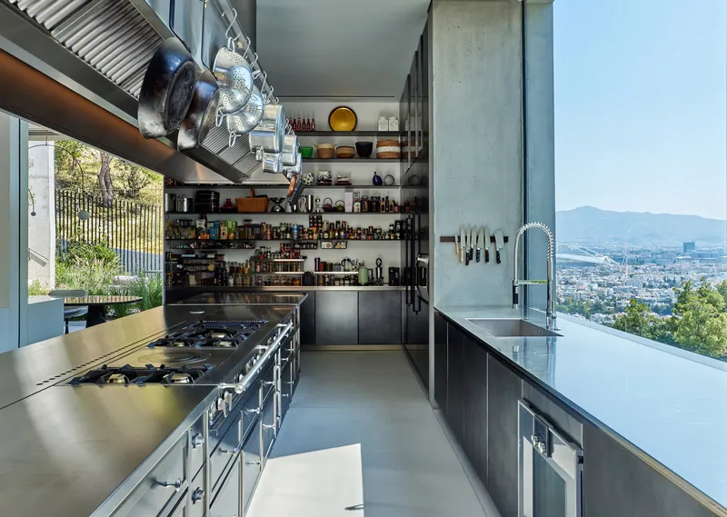 Galley kitchen with steel counters and open shelving overlooking the hillside through floor-to-ceiling glass