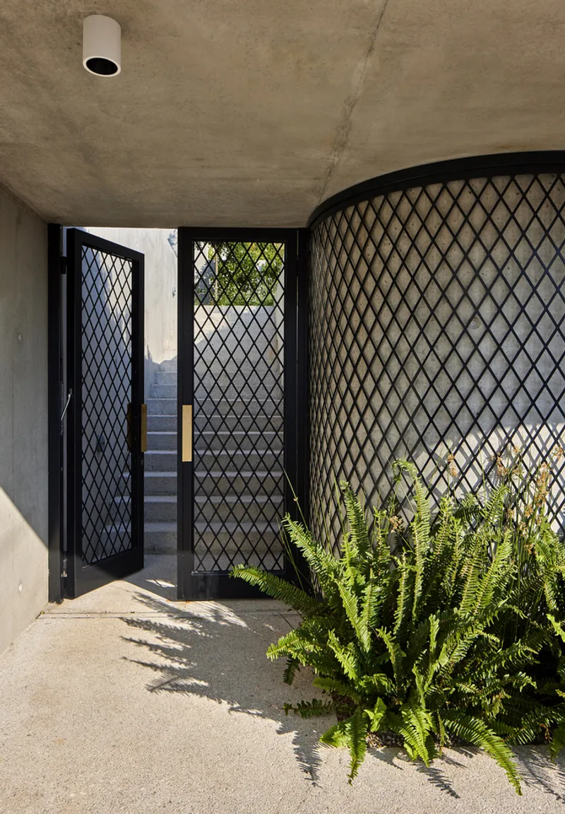 Covered entry with concrete ceiling and black diamond mesh screen doors beside planted ferns in dappled sunlight