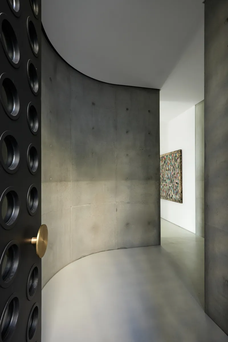 Curved concrete corridor with brass door handle and circular perforations in the wall catching soft daylight