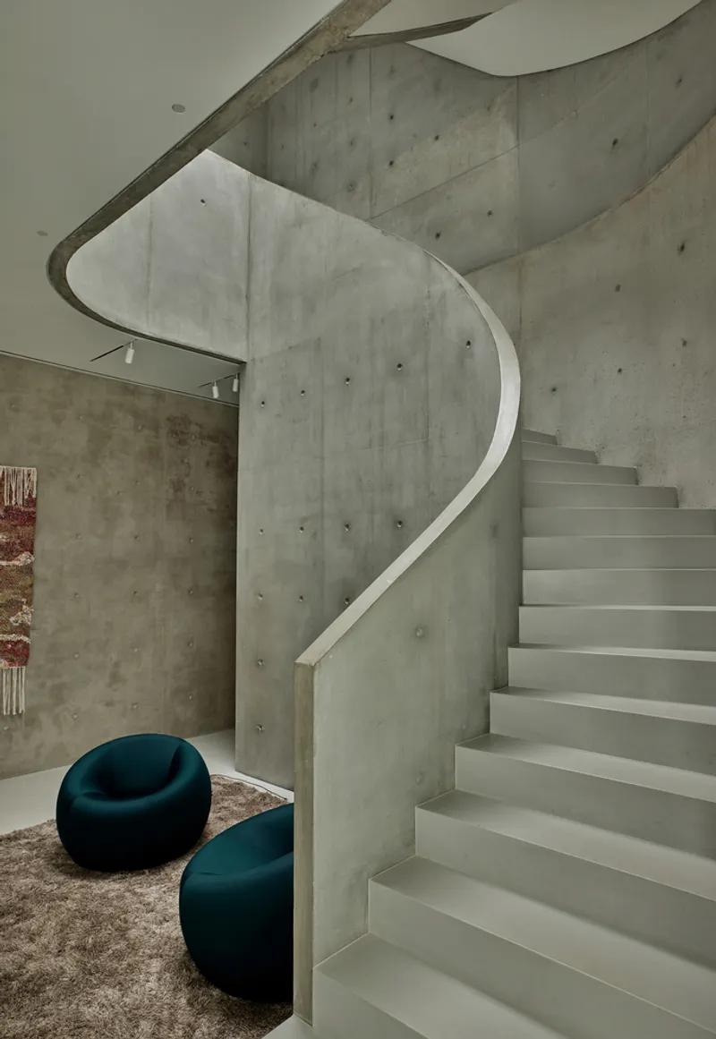 Spiraling concrete staircase with sculptural soffit and two upholstered chairs beneath the curved volume