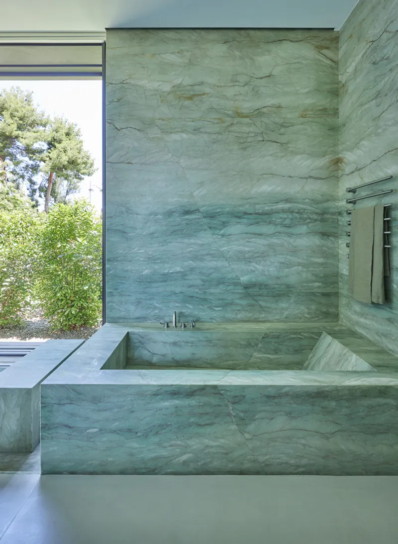 Sunken bathtub clad in book-matched green marble with a window framing trees outside