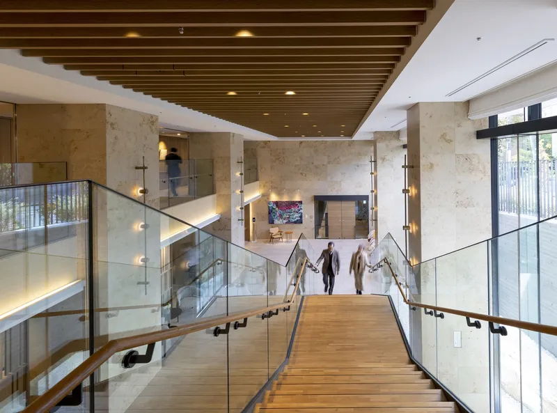 Timber and glass staircase descending through a multi-level lobby with limestone walls and residents walking