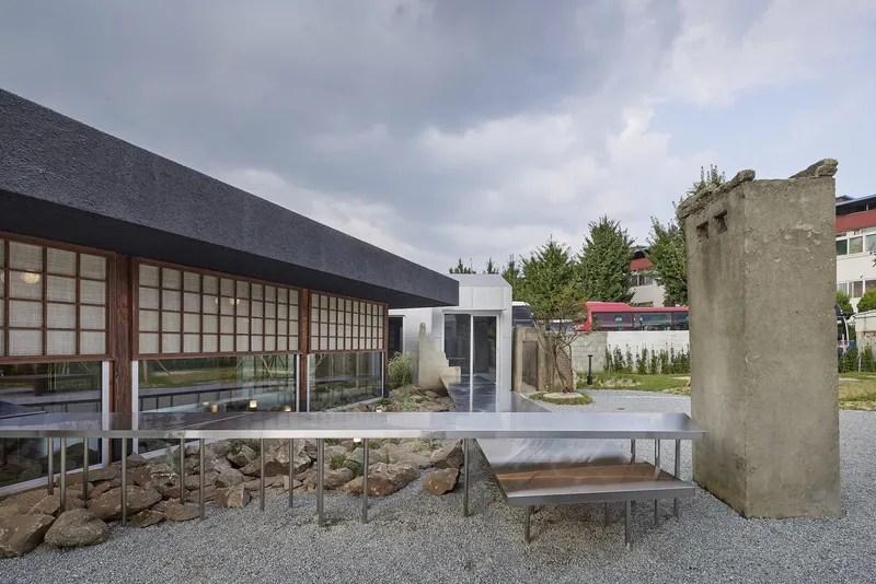 Gravel courtyard with elevated steel table and weathered concrete ruin under an overcast sky