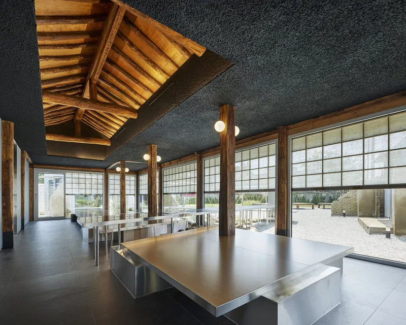Interior hall with timber roof structure, translucent grid windows, and stainless steel tables on dark flooring