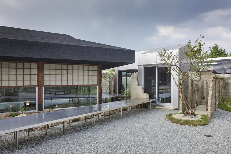Gravel courtyard showing the pavilion with gridded facade, elevated steel walkway, and planted tree at dusk