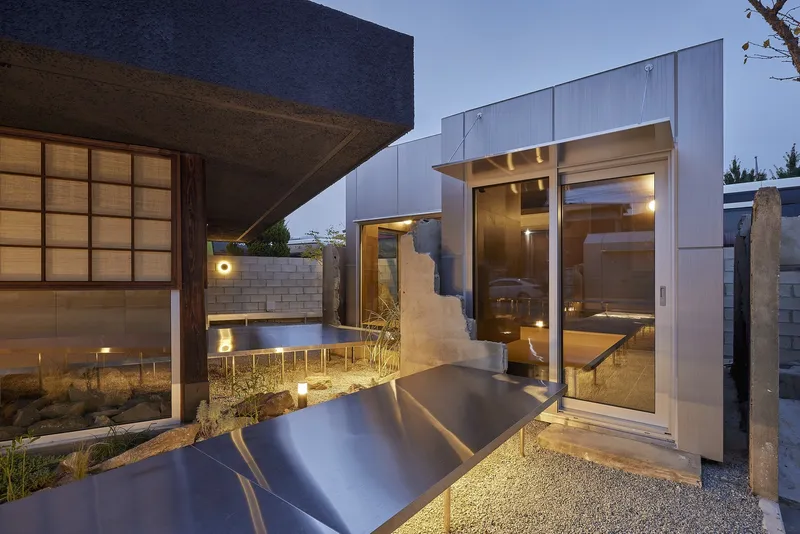 Courtyard with gravel ground and metal-clad volumes beside gridded sliding doors at dusk