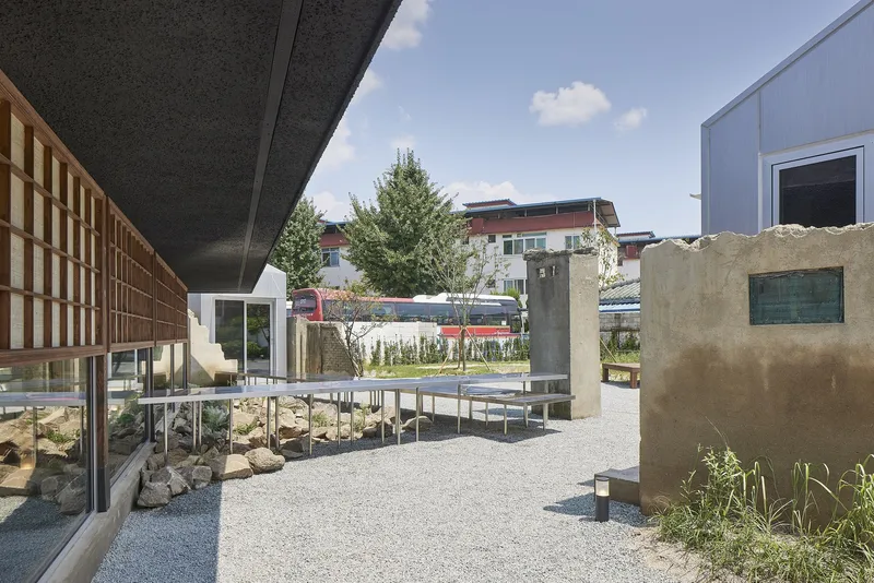 Gravel courtyard with metal benches, planted beds, and gridded timber screens under a blue sky
