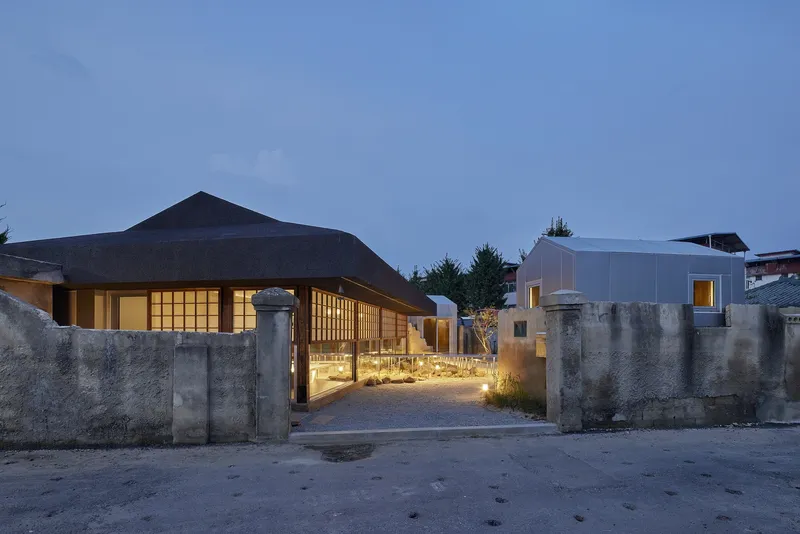 Entry courtyard with gravel path, concrete walls, and illuminated timber-framed pavilions at dusk