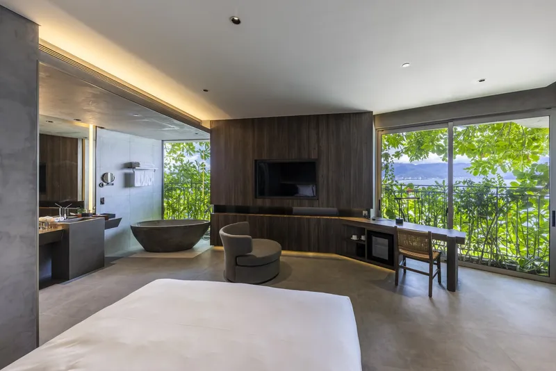 Hotel room with walnut wall paneling, freestanding tub, and full-height glazing overlooking planted balcony