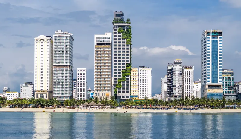 Coastal skyline showing residential towers with green facade rising above palm-lined beach under cloudy sky