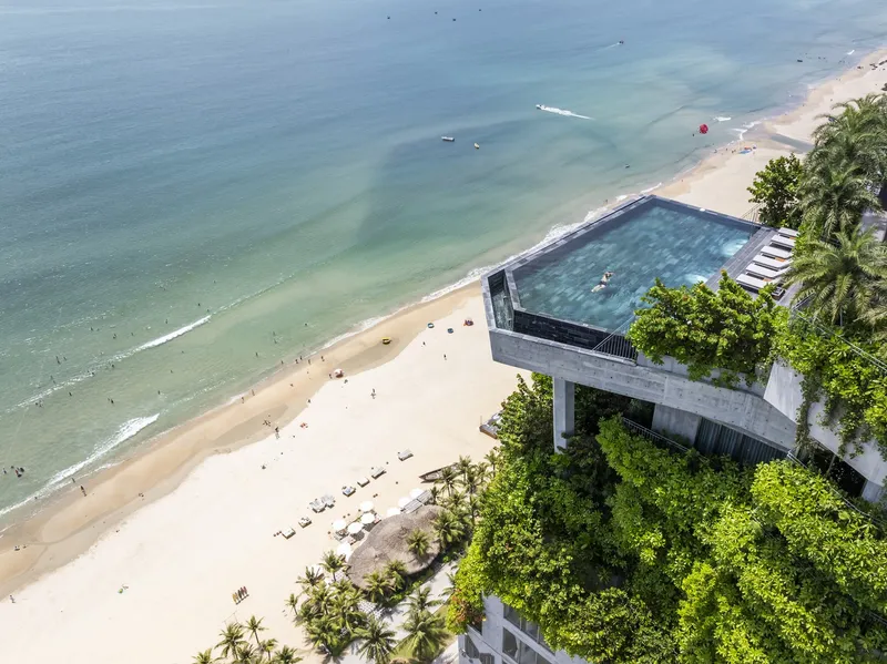Aerial view of the cantilevered rooftop pool over the beach with palm trees and calm water