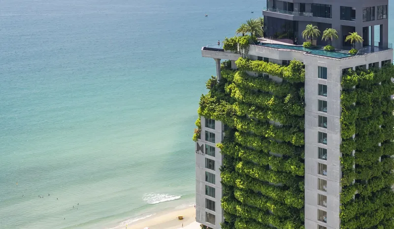 Aerial view of tower facade with cascading planted terraces rising above turquoise coastal waters