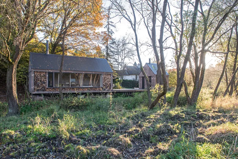 Two gabled timber structures with corrugated metal roofs and stacked firewood on the facade in an overgrown meadow