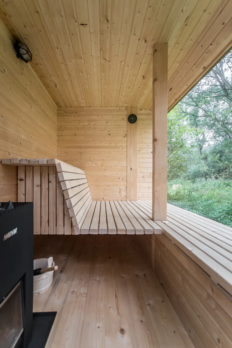 Timber sauna interior with curved bench slats, tongue and groove ceiling, and wood-burning stove under trees