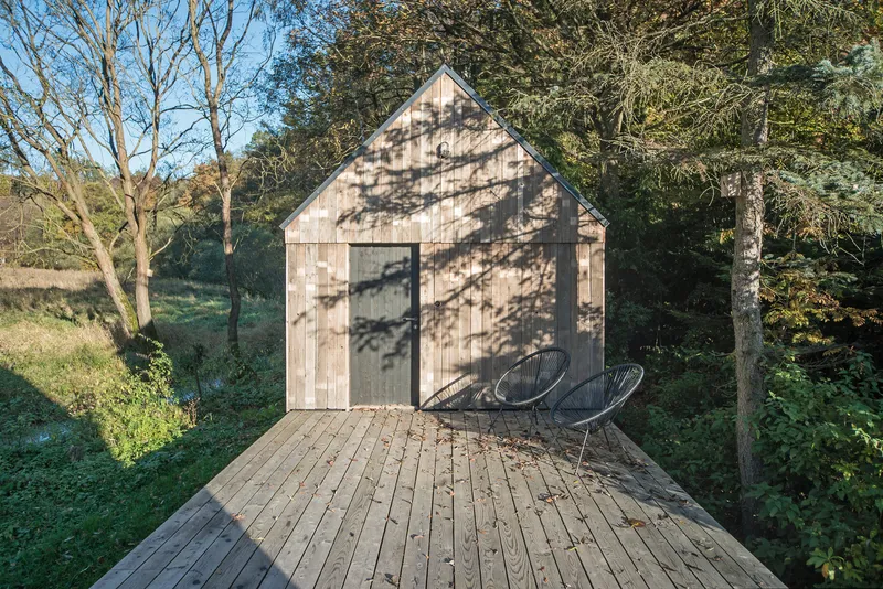 Cedar-shingled gable facade with glazed corner and dappled tree shadows on a raised timber deck