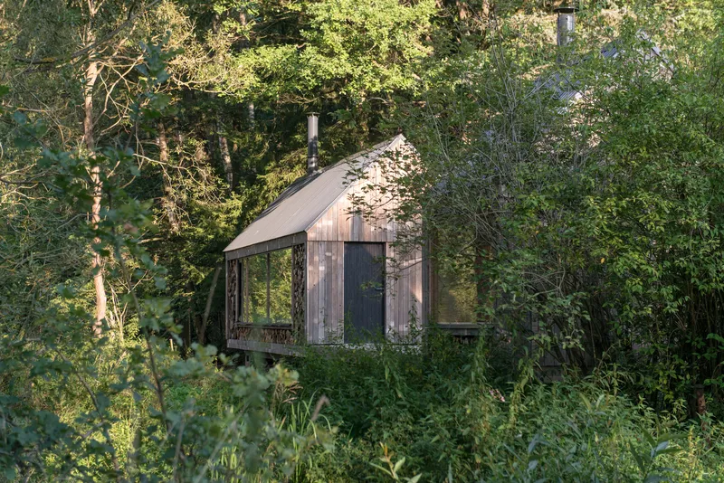Weathered timber cabin with metal roof and stacked firewood gable surrounded by dense summer vegetation