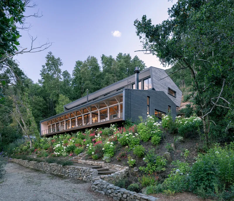 Dark timber-clad house on a sloped garden with stone retaining walls and flowering shrubs at dusk
