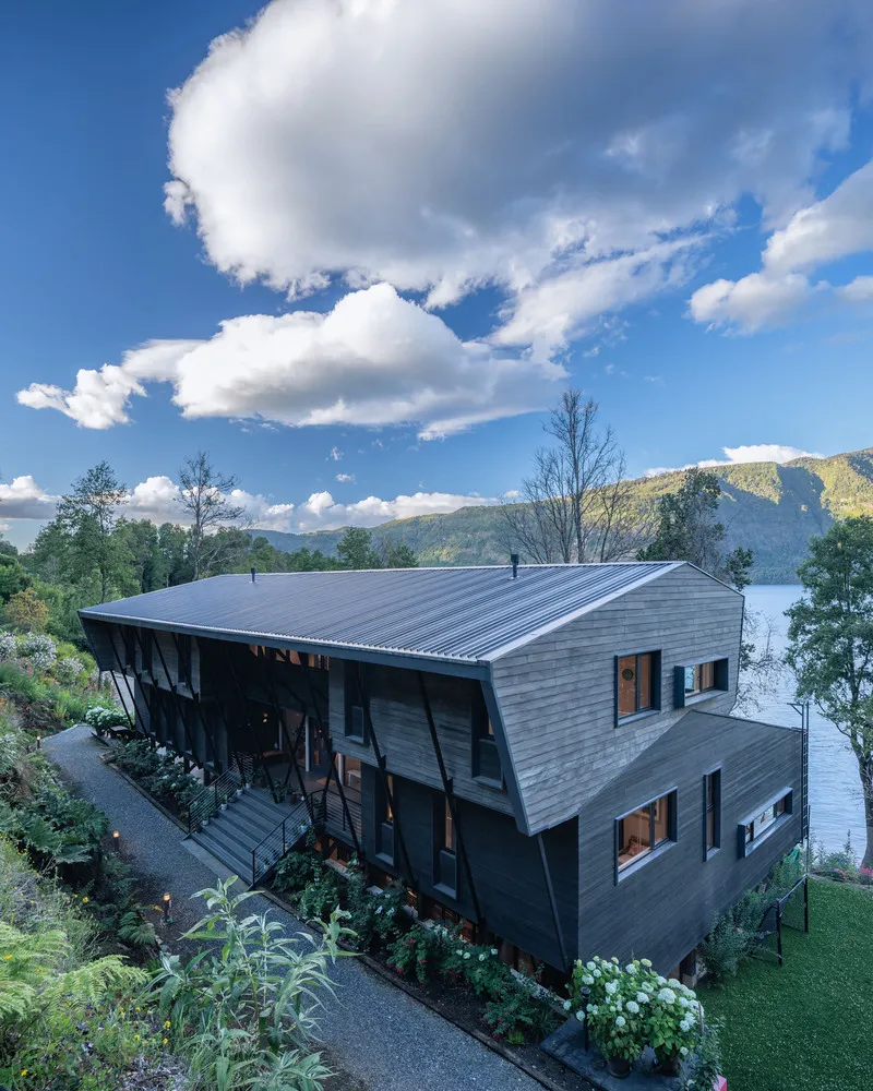 Elevated view of the angular wood-shingled residence with lake and mountains visible beyond the trees