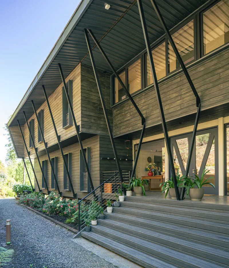Covered deck with timber steps and diagonal black steel bracing along the facade at sunrise