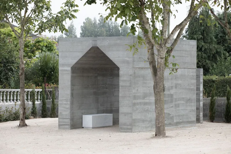 Board-formed concrete chapel volume with gable cutout among deciduous trees and gravel courtyard