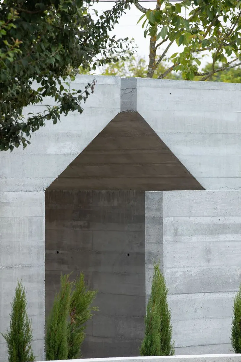 Boardformed concrete facade with triangular gable entry flanked by slender cypress plantings