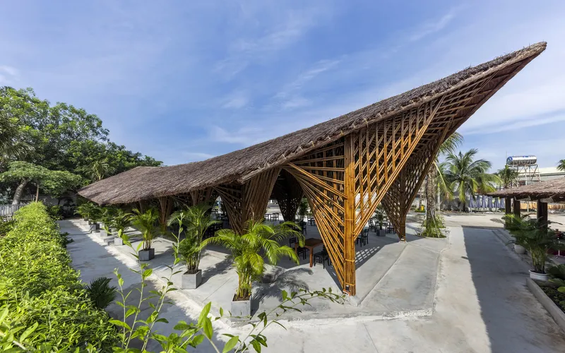 Thatched bamboo pavilion with sweeping angled roof and exposed lattice columns beside palm trees under blue sky